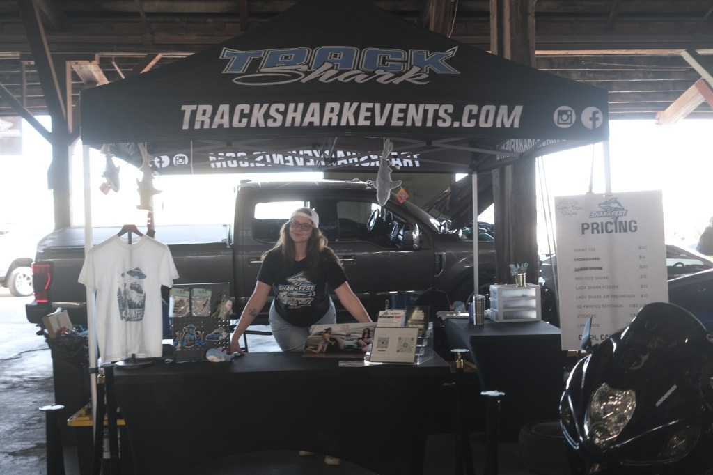 A vendor booth at an event featuring a woman smiling in a black t-shirt and cap, standing behind a table with merchandise and a pricing sign. A truck is visible in the background under a large tent displaying the Track Shark logo.