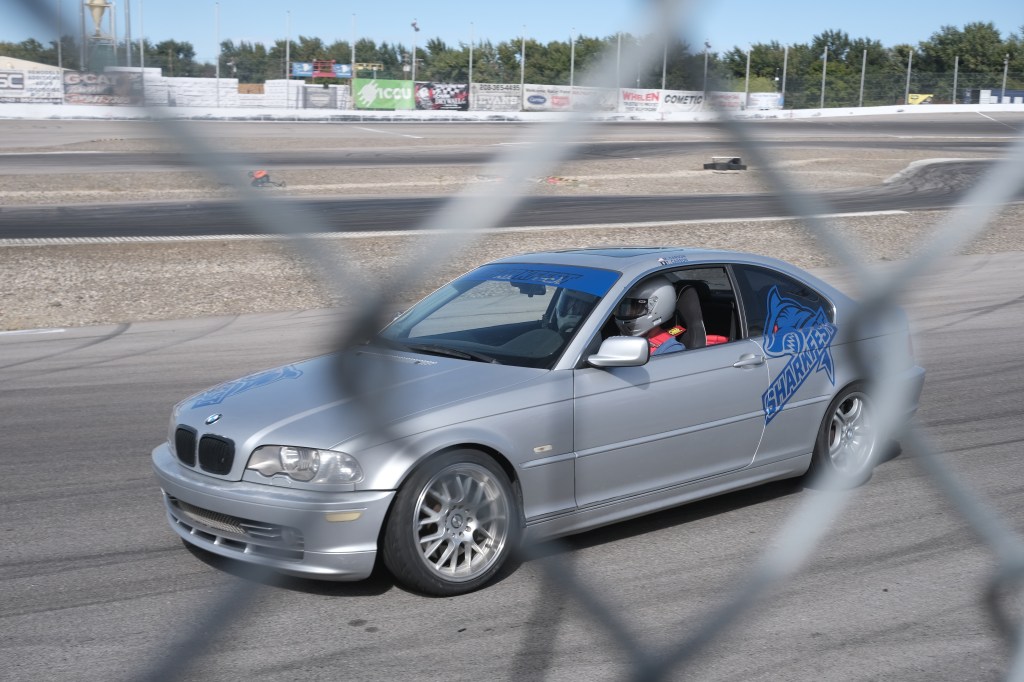 A silver BMW car with a shark logo on the side, driving on a racetrack behind a fence, with a driver wearing a helmet.