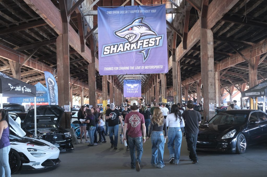 A crowded automotive event at SharkFest with banners and booths under a wooden structure, showcasing various cars and attendees walking around.
