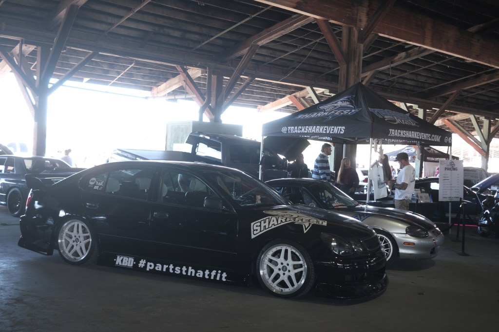 A black modified car with custom wheels parked indoors under a wooden structure, alongside other cars and a tent displaying event details.