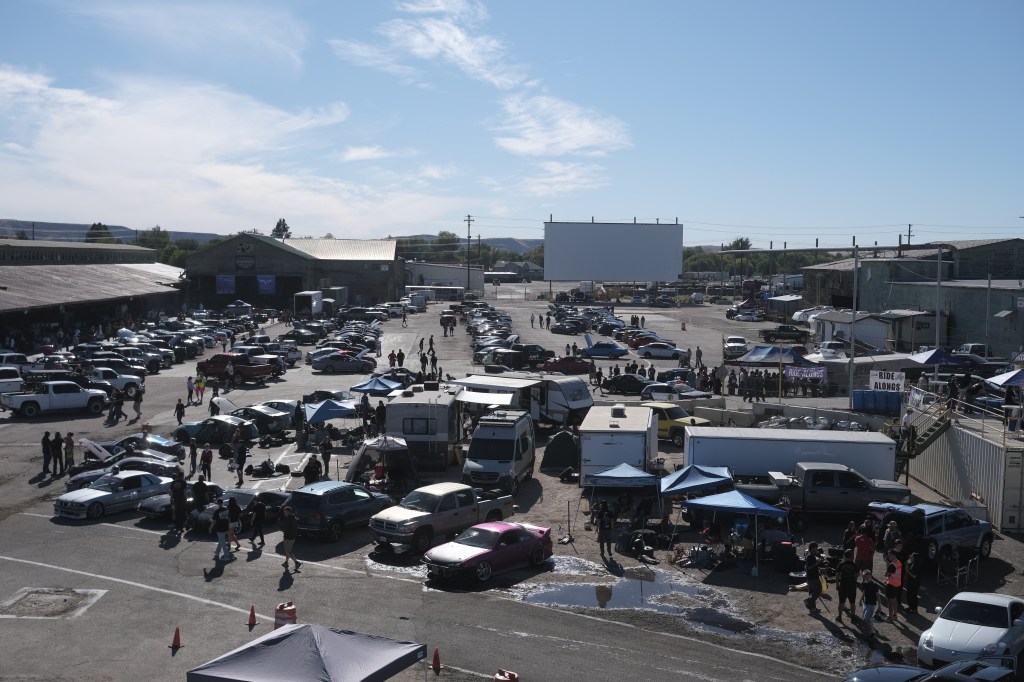Aerial view of a busy outdoor car meet, featuring numerous parked vehicles, tents, and people gathered in a large open area under clear blue skies.