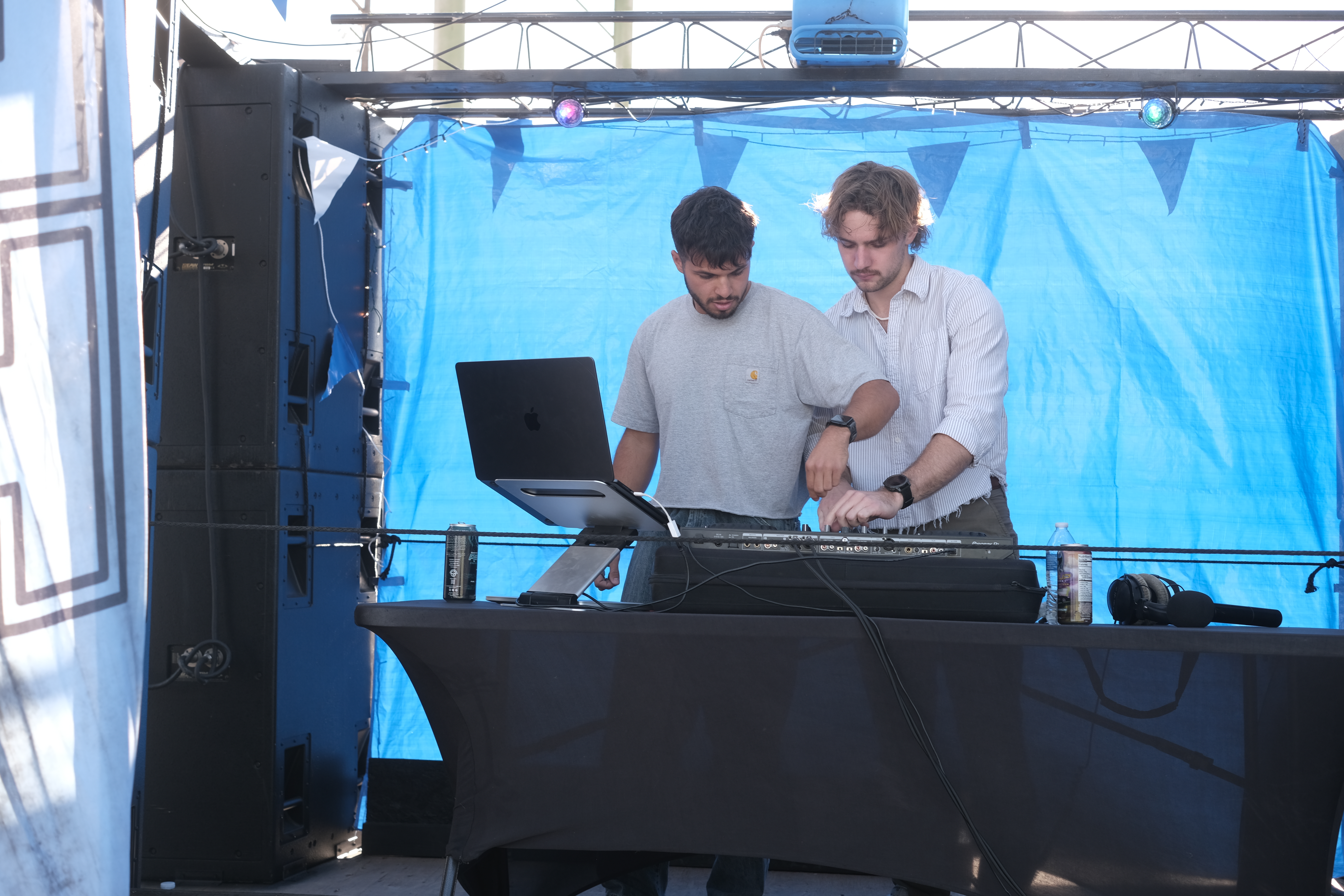 Two men working together behind a DJ booth with a laptop and equipment, set against a blue backdrop.