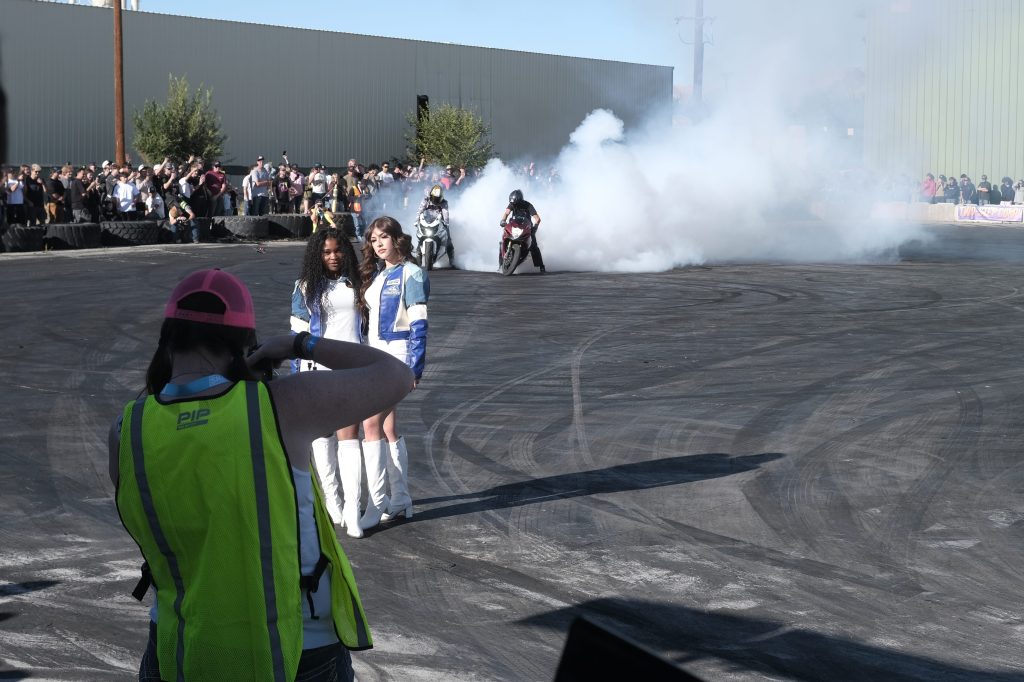 A crowd watches a motorcycle performing a burnout, creating smoke on a racetrack. Two women in racing outfits stand in the foreground, while a person in a safety vest is taking a photo.