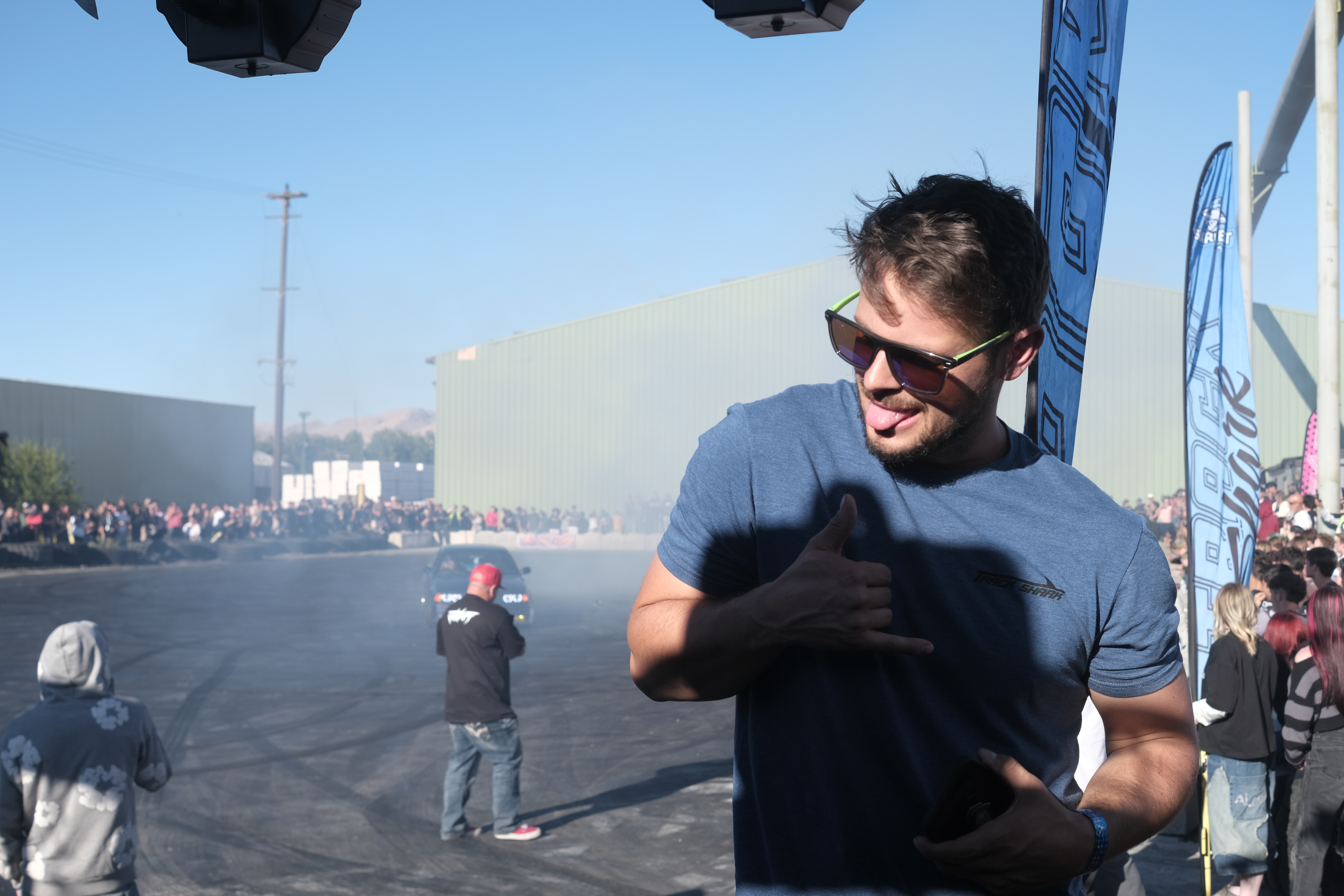 A person making a shaka sign while standing at an outdoor event with a crowd in the background and smoke from a car drifting.